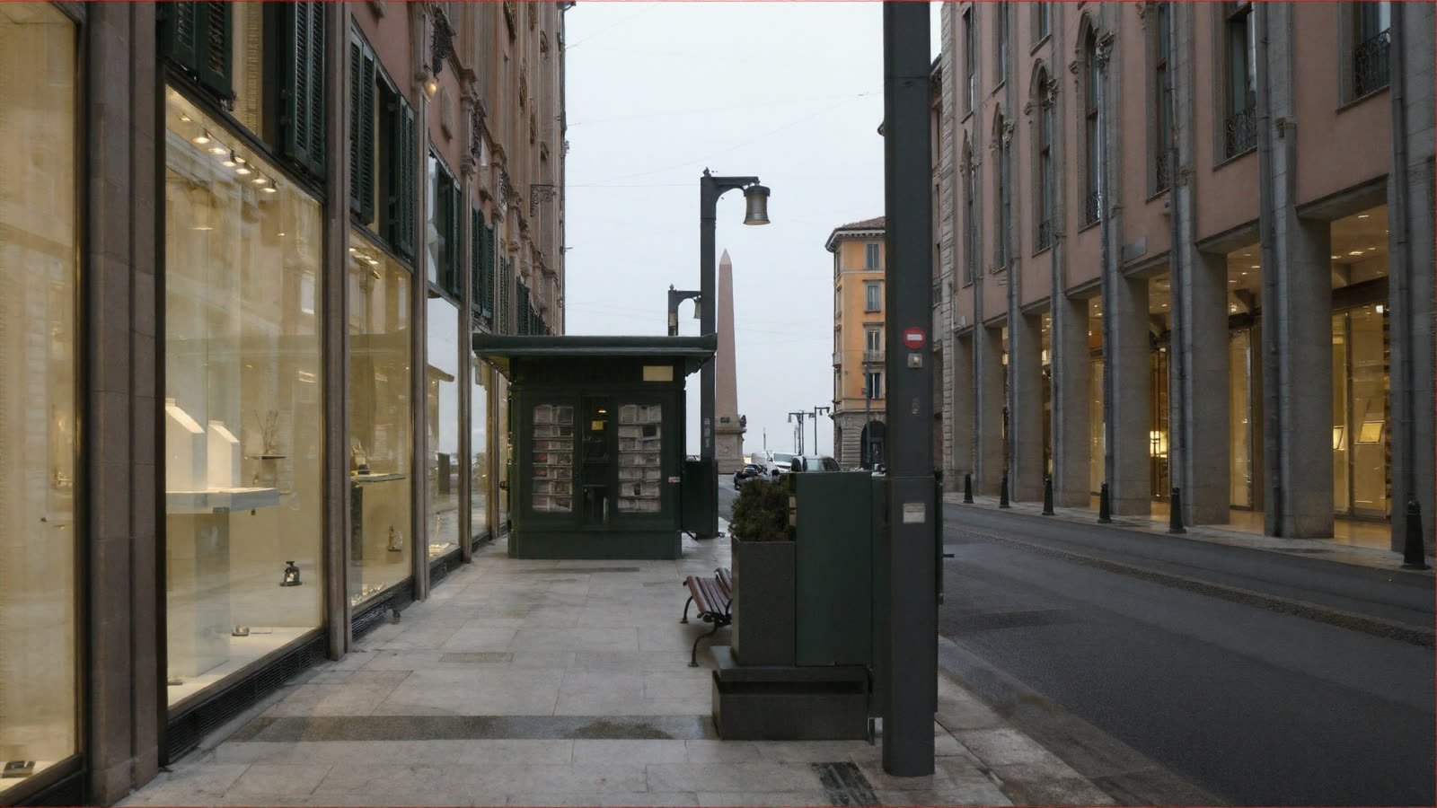 Looking north along the street: lit shopfronts on the left, a green newspaper kiosk mid-frame, an arcade of piers on the right, an obelisk in the distance under flat grey cloud