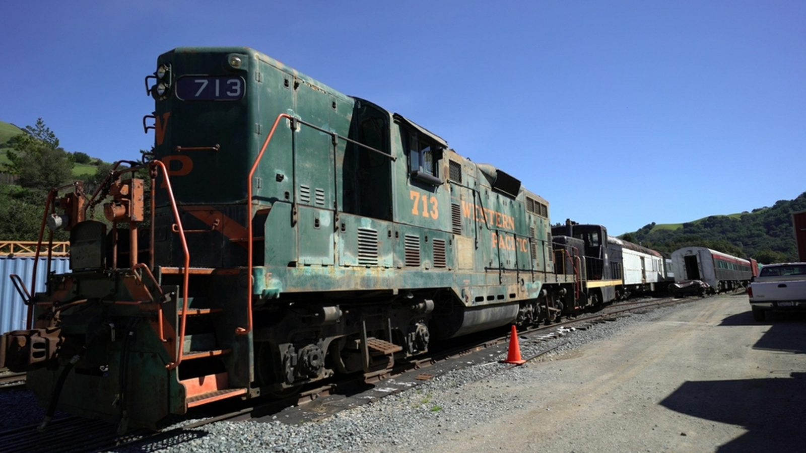A photograph of a preserved green diesel locomotive numbered 713 standing in a railway yard under a blue sky, with an orange traffic cone beside the track, passenger carriages behind it and a corrugated shed to the left.