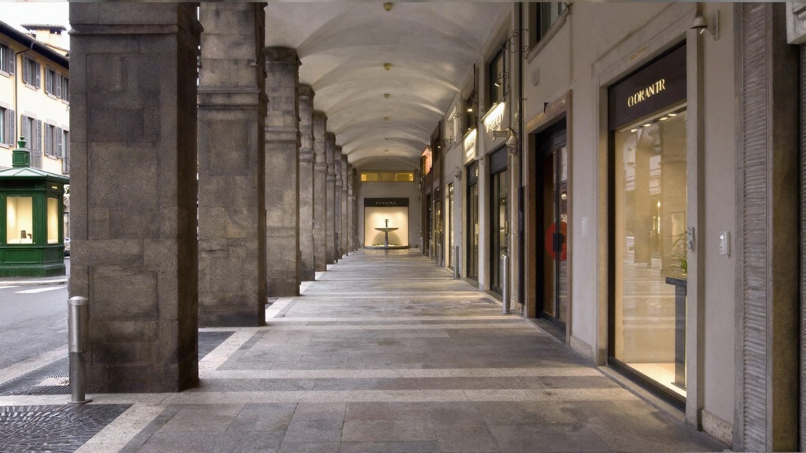 Inside a stone arcade with vaulted ceiling and lit shopfronts