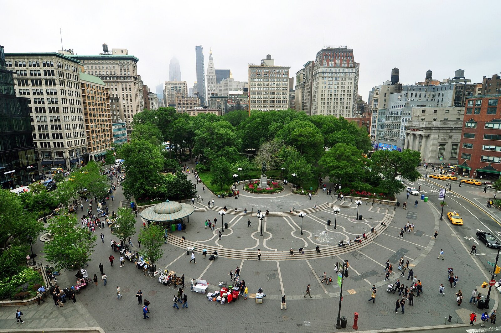 A photograph of the real Union Square looking north from above the south plaza: the circular paving ring, the green-domed pavilion, the equestrian statue in its planted circle, dense green plane trees, pre-war buildings around the square and the midtown skyline behind.