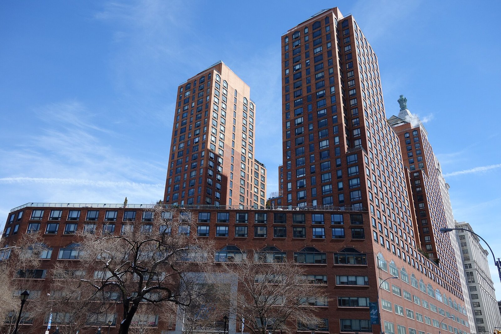 A photograph of the real Zeckendorf Towers seen from below against a blue sky: dark red-brown brick towers with paler buff brick panels, dark tinted windows in a dense grid, stepping back from a broad brick base block that carries a glass balustrade along its roof.