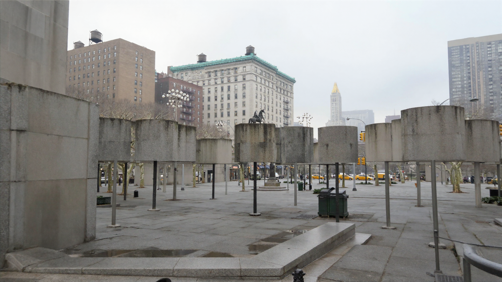 A photorealistic view of a New York square in which the trees have been rendered as grey concrete cylinders mounted on thin steel poles, standing in rows across a granite plaza, with genuine pre-war buildings and a bronze equestrian statue behind them.