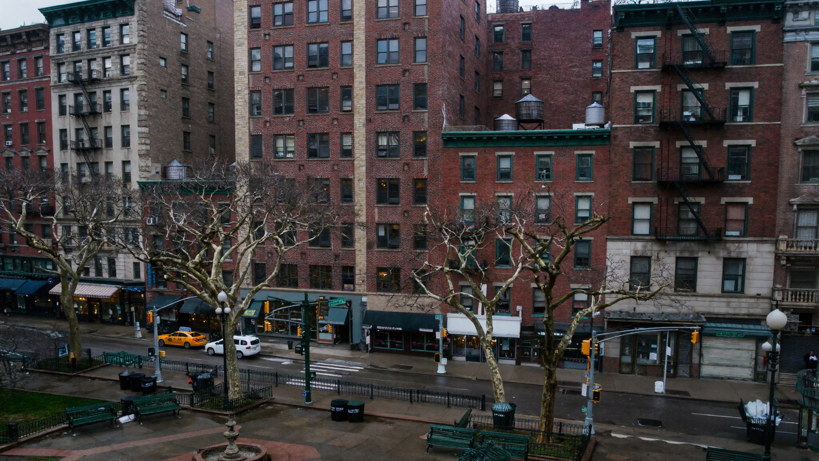A photorealistic overcast view of the east side of Union Square: red brick buildings eight to ten storeys tall with black iron fire escapes and wooden water tanks on the roofs, shopfronts under dark green awnings at street level, bare plane trees, green cast-iron benches and railings, a yellow taxi and a white car on the wet street.
